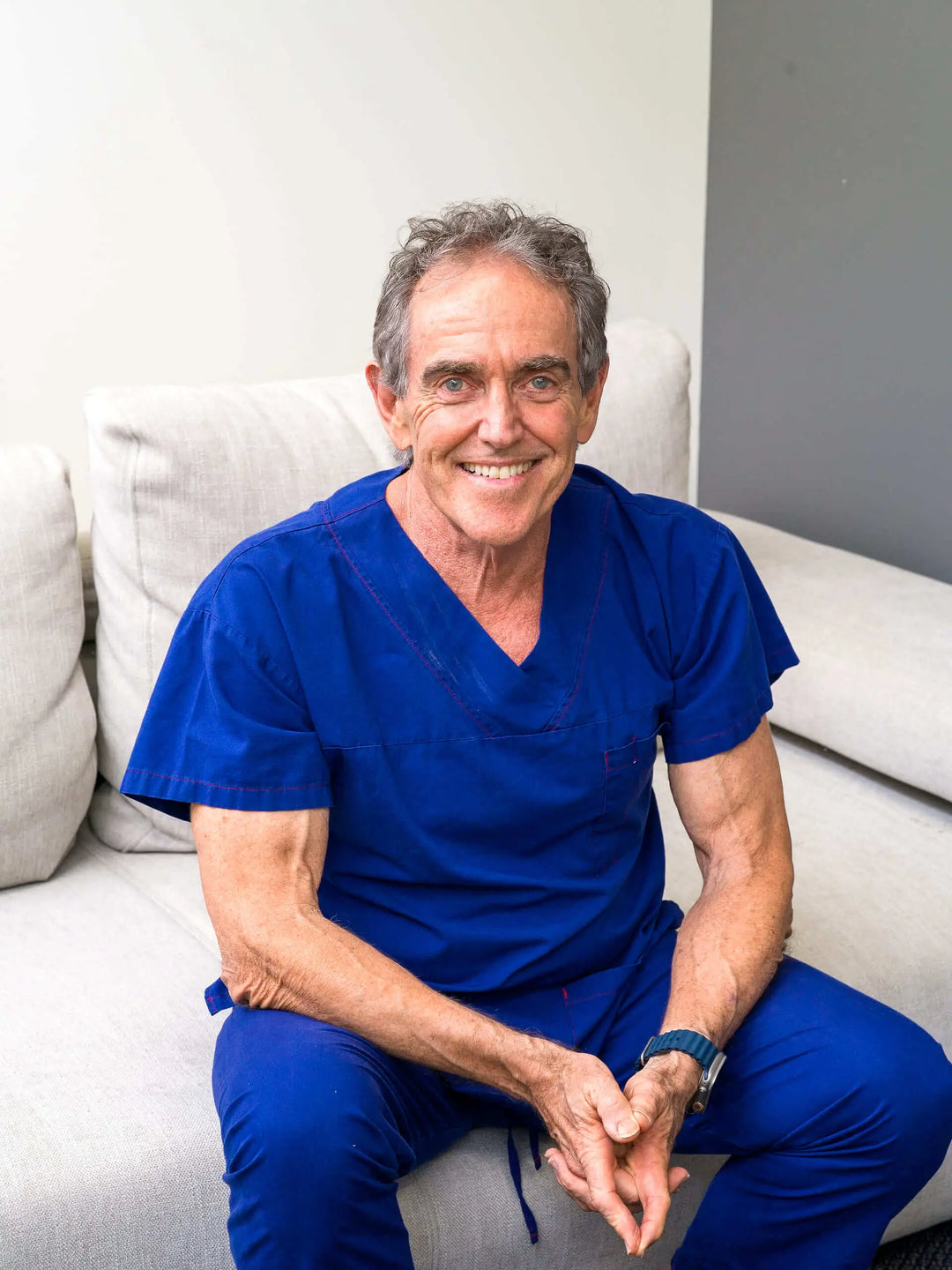 Doctor Mark Doyle smiling in blue medical scrubs while seated on a sofa