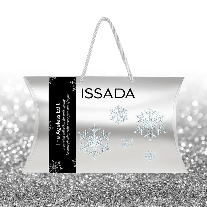 Front view of Issada The Ageless Edit festive silver skincare gift bag with black side label, snowflake design, and white rope handles, displayed on a silver glitter background.
