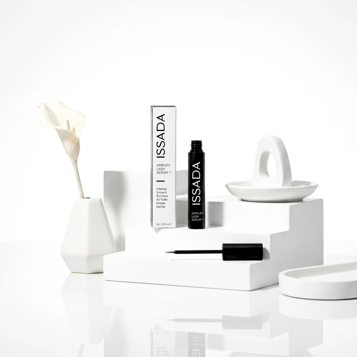 ISSADA Amplify Lash Serum with packaging and applicator styled on white geometric props beside a calla lily.