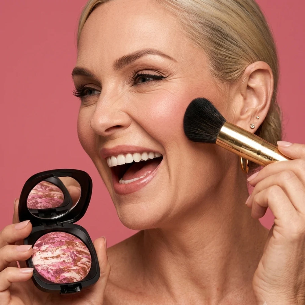 Smiling woman applying Mineral Baked Blush Raspberry Ripple with a brush while holding an open compact against a pink background.