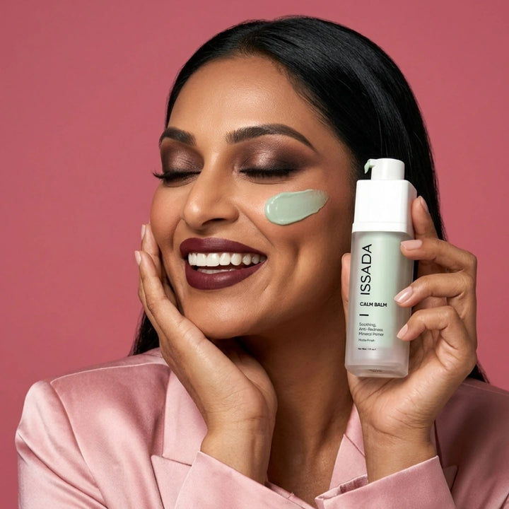 Smiling woman holding ISSADA Calm Balm Primer with a mint-green swatch on her cheek against a pink background.