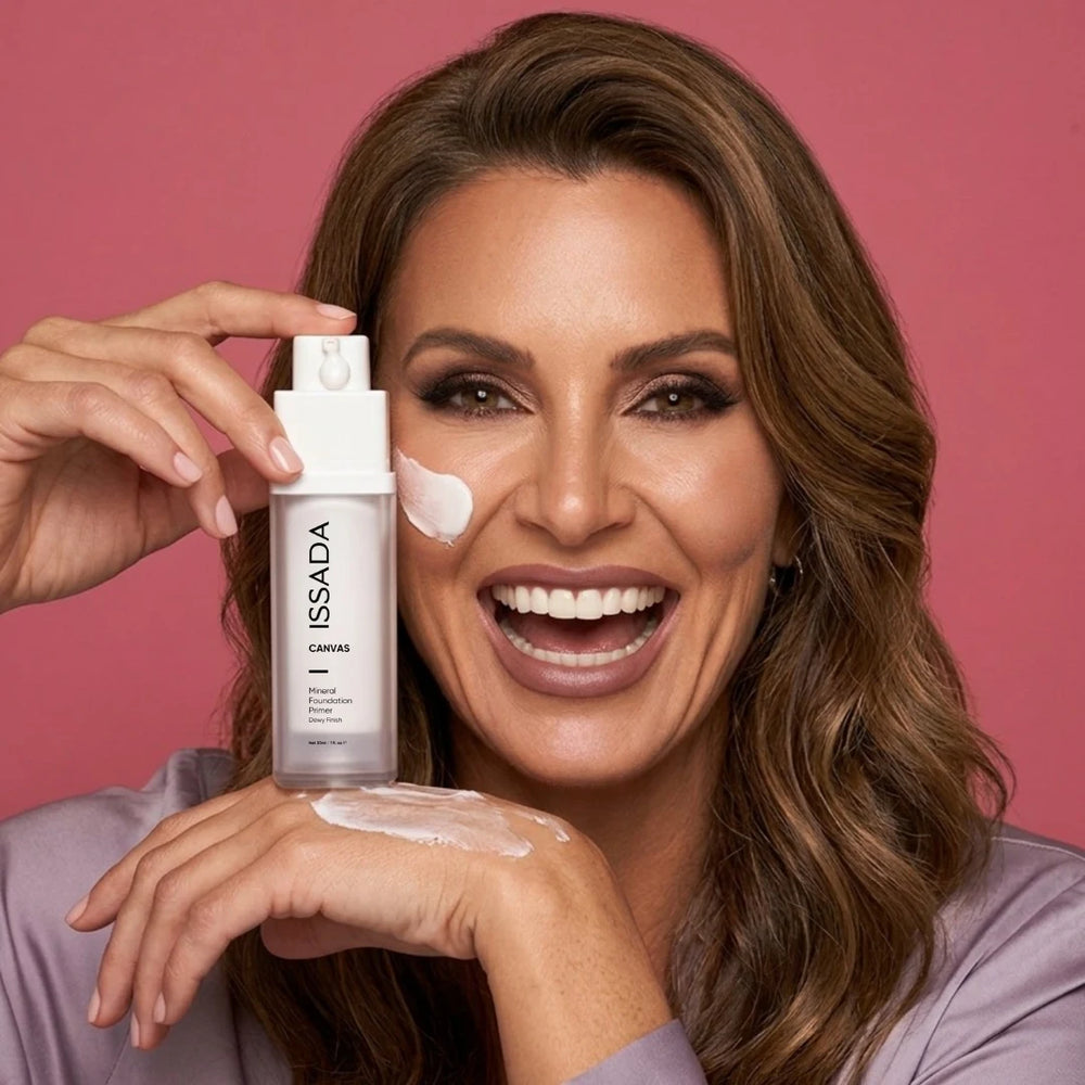 Smiling woman holding ISSADA Canvas Primer with white primer swatches on her cheek and hand against a pink background.