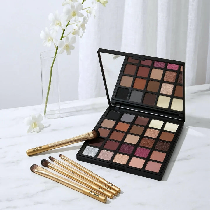 Open ISSADA Clay & Ember eyeshadow palette with warm neutral shades and gold makeup brushes beside white orchids.