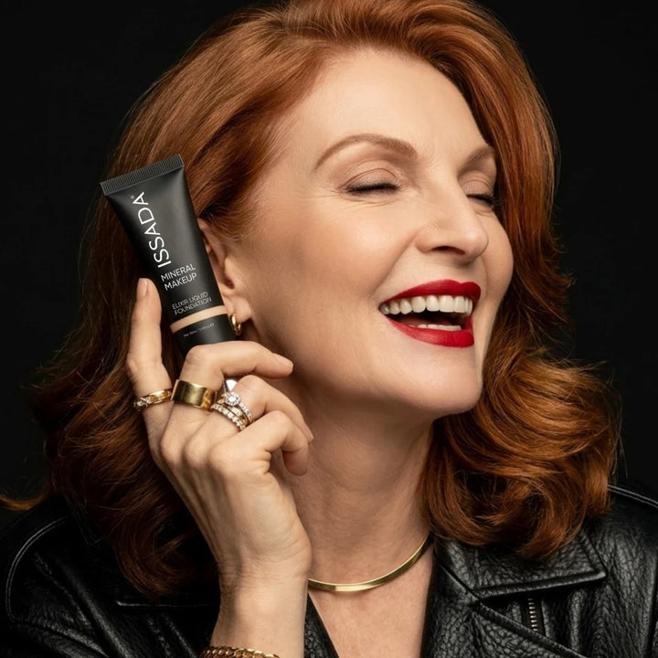 Smiling red-haired woman holding ISSADA Elixir Liquid Foundation against a black background with bold red lipstick.