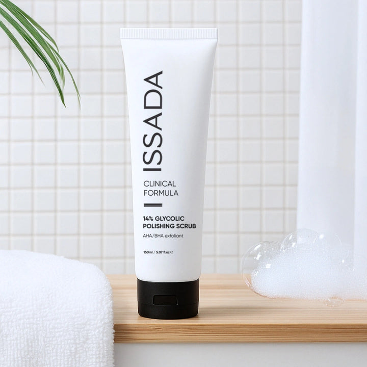 ISSADA 14% Glycolic Polishing Scrub tube on a bathroom shelf beside a white towel and foamy bubbles.