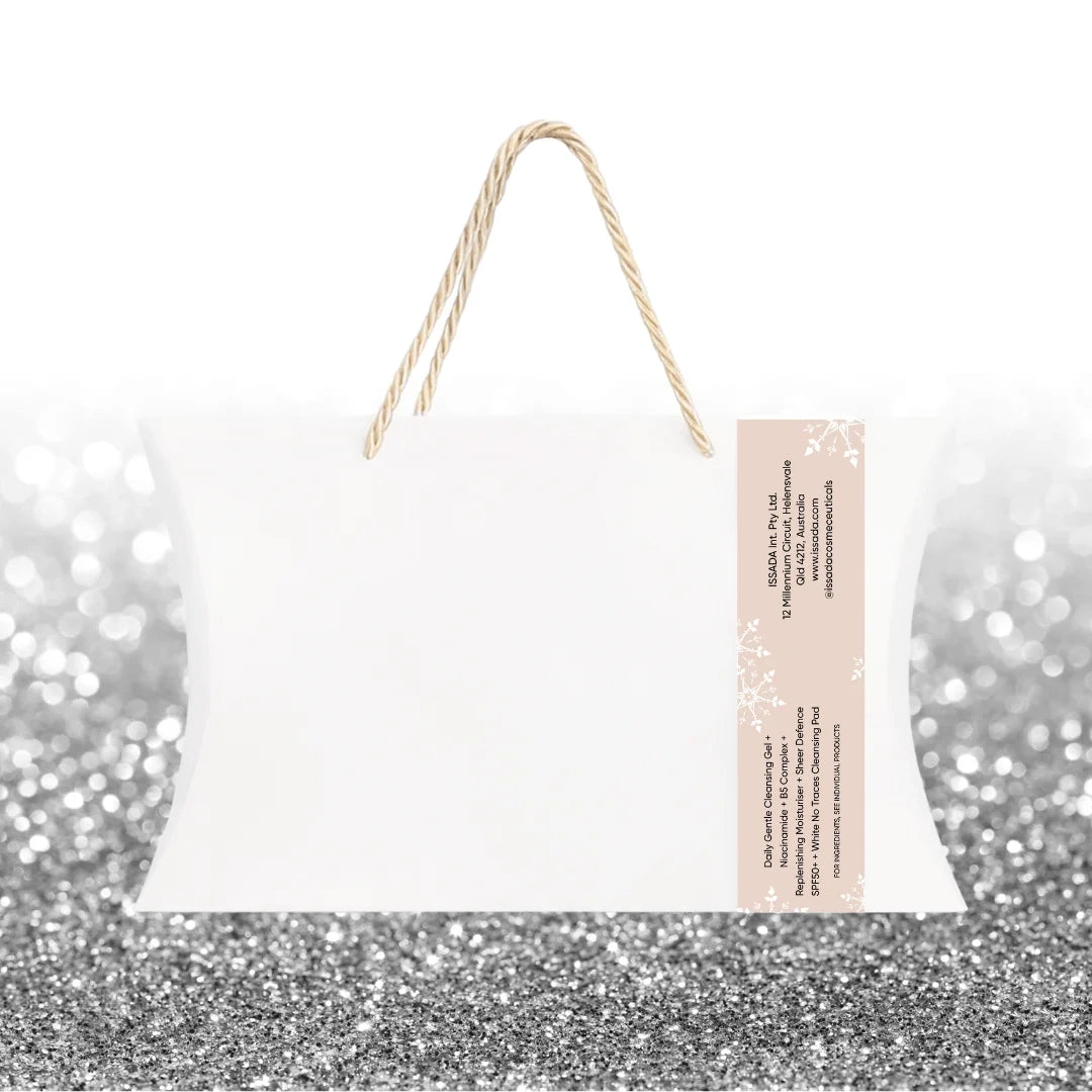 Back view of Issada Fresh Faced Festive Collection white holiday gift bag with rope handles and beige label text, displayed on a silver glitter background.