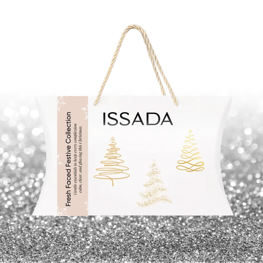 Issada Fresh Faced Festive Collection closed white gift bag with gold Christmas tree designs and rope handles on a silver glitter background.