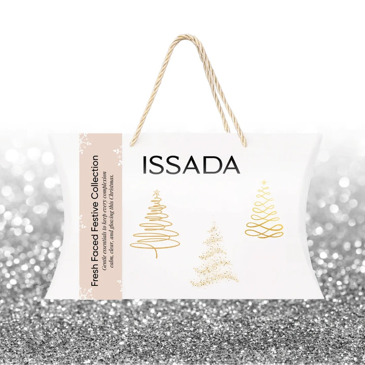 Issada Fresh Faced Festive Collection closed white gift bag with gold Christmas tree designs and rope handles on a silver glitter background.