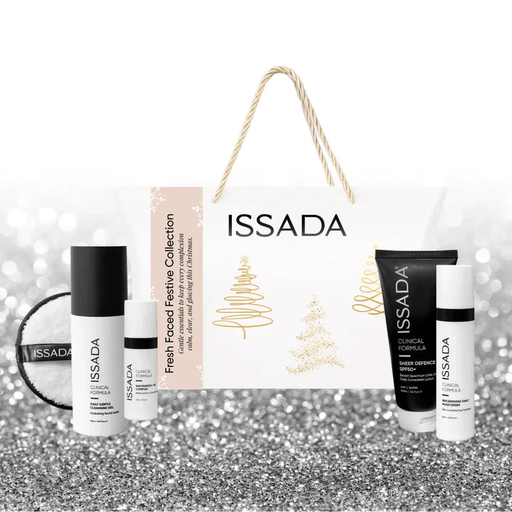 Issada Fresh Faced Festive Collection white holiday gift bag with gold accents, featuring skincare products including cleanser, SPF, serum, and cleansing pad on a glitter background.