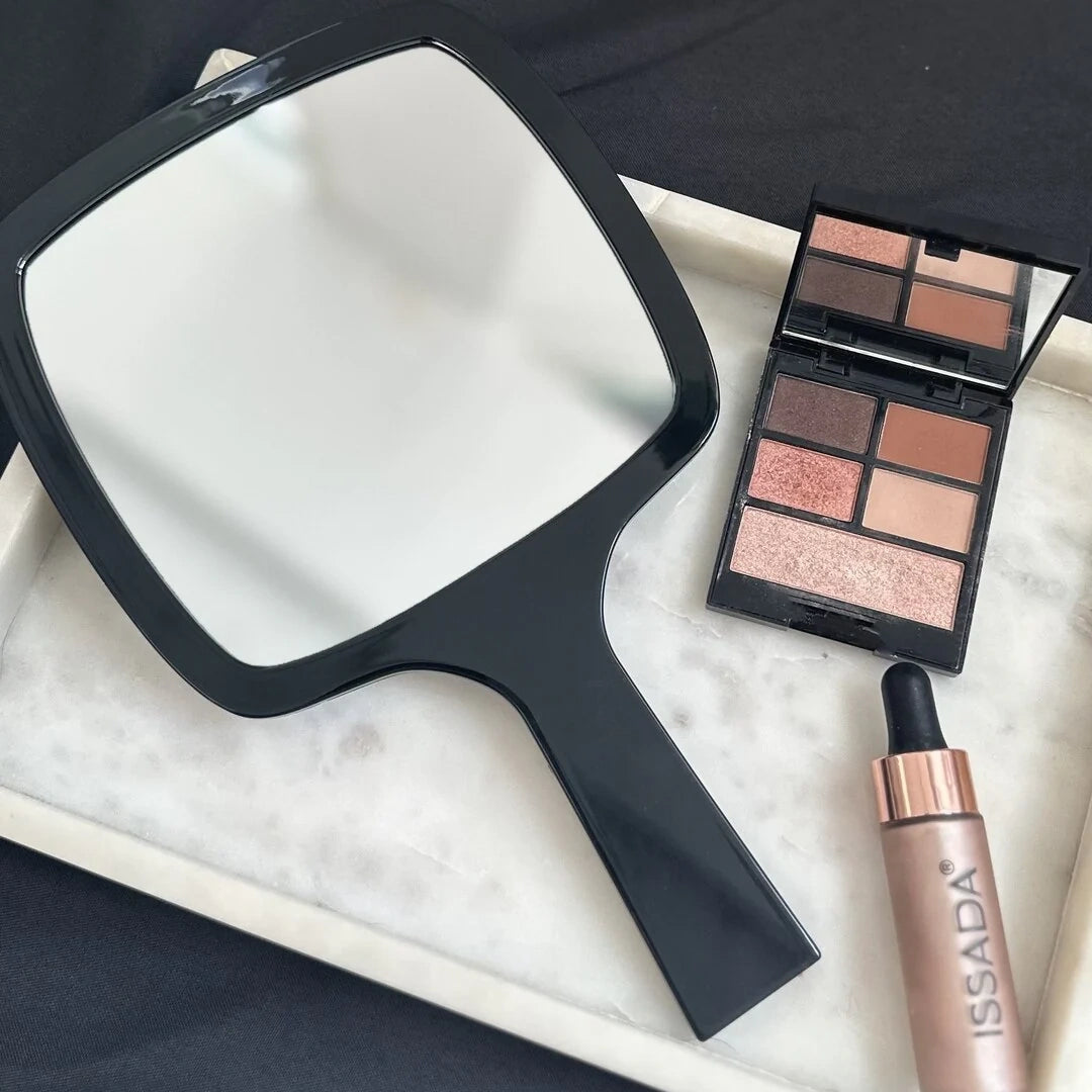 Issada black handheld mirror on marble tray with eyeshadow palette and liquid highlighter for professional makeup application.
