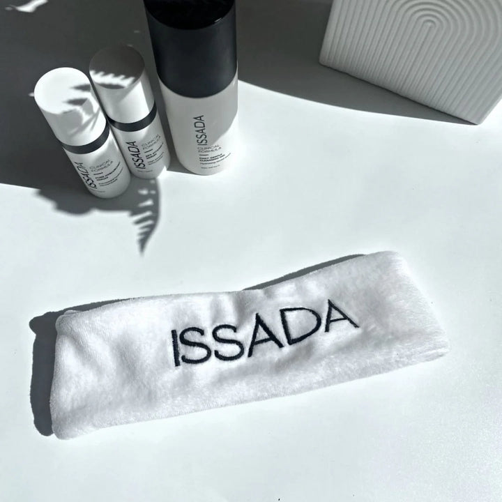 White ISSADA cosmetic headband with embroidered logo displayed beside Issada skincare products on a white surface