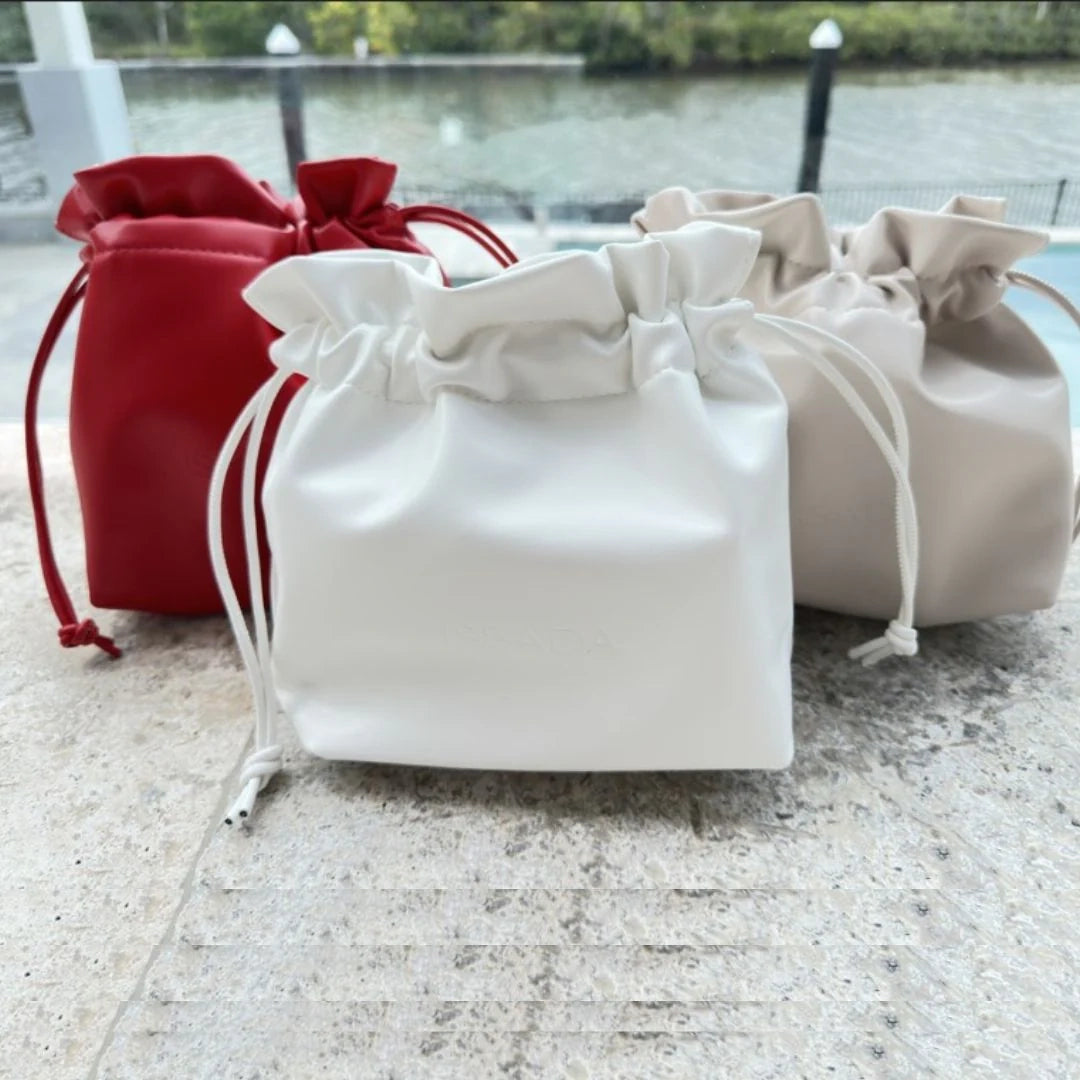 Red, white, and nude Issada drawstring cosmetic bags displayed outdoors with a river view in the background.