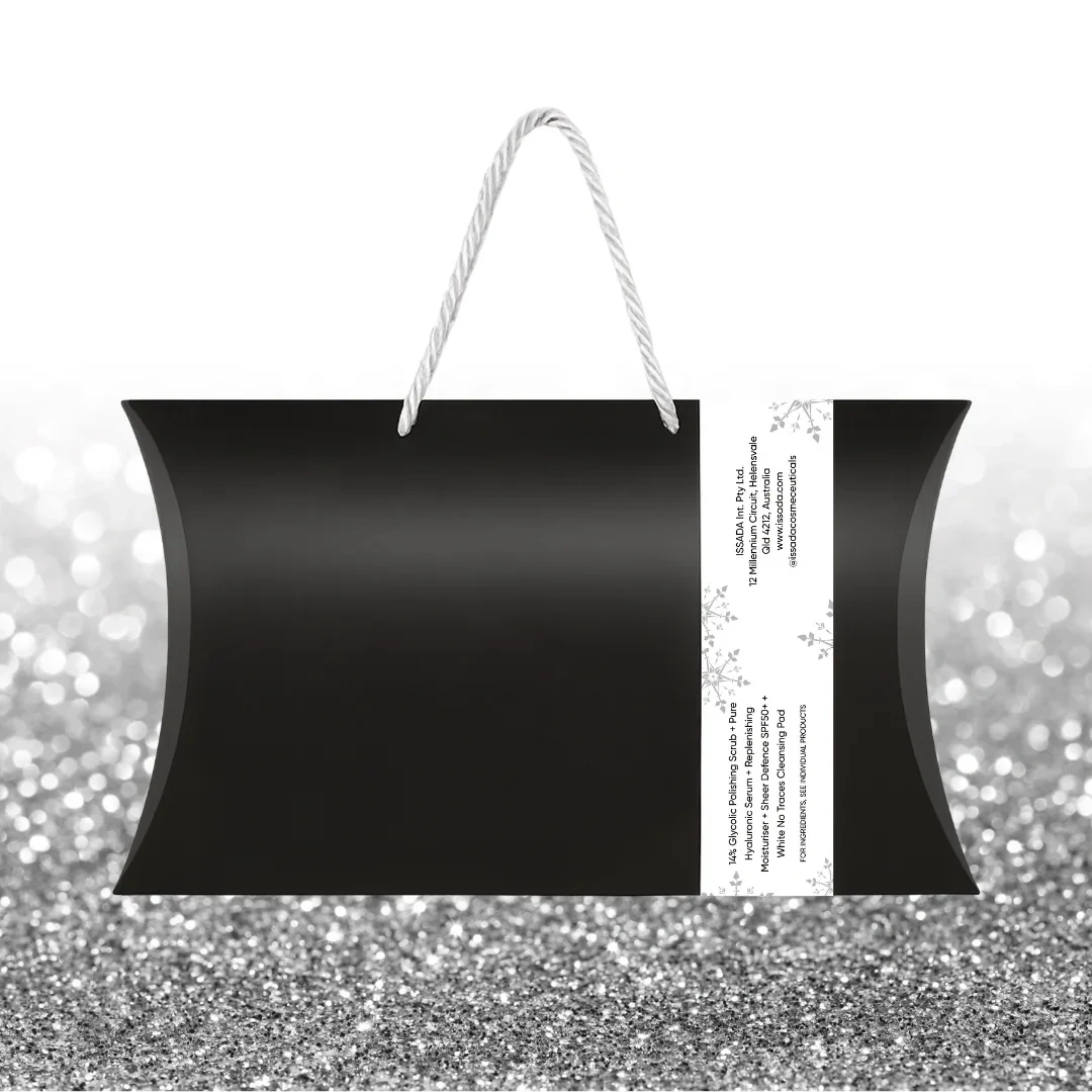 Back view of Issada Merry & Bright Skin Pack black gift bag with white rope handles and side product information label, displayed on a silver glitter background.