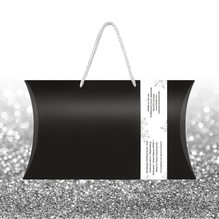 Back view of Issada Merry & Bright Skin Pack black gift bag with white rope handles and side product information label, displayed on a silver glitter background.