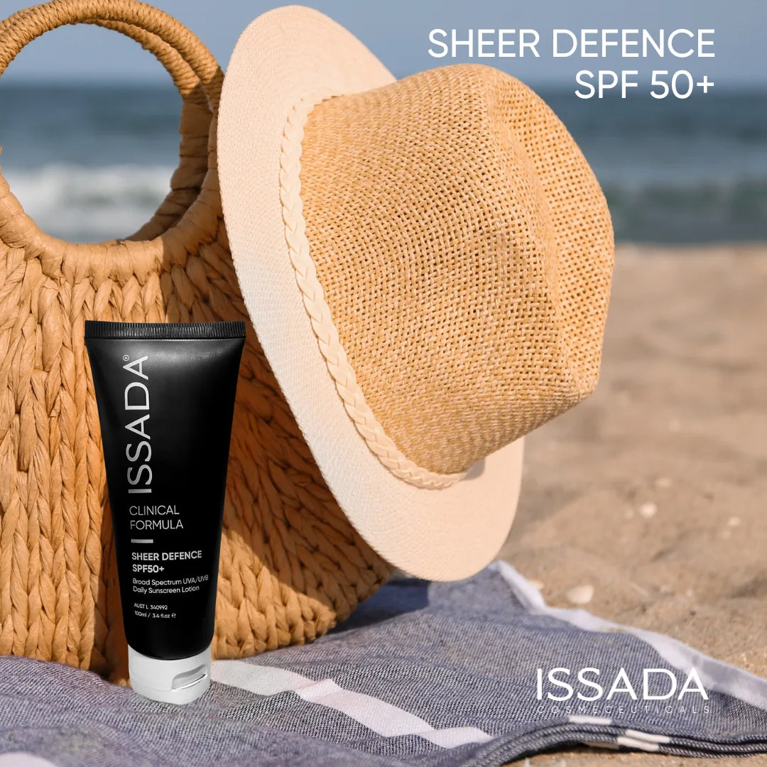 Issada Sheer Defence SPF50+ sunscreen placed on the beach beside a woven hat and bag, highlighting sun protection for outdoor use.