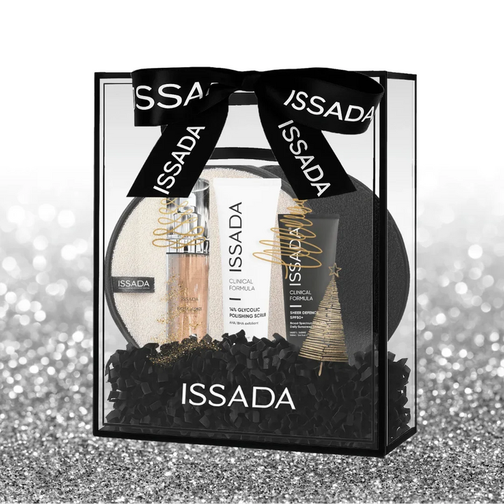 Issada Shimmer Into Summer gift set featuring Hydraglow serum, glycolic polishing scrub, sheer defence SPF50, and cleansing pad in a clear gift box with black ribbon and festive accents on a glitter background.