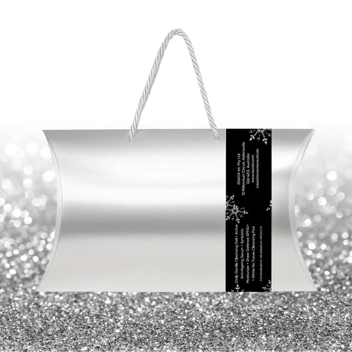 Back view of Issada The Ageless Edit festive silver skincare gift bag with white rope handles, black side label, and product details printed on the label, placed on a silver glitter background.