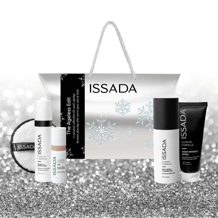 Issada The Ageless Edit festive silver gift bag with white rope handles and snowflake design, displayed with four clinical skincare products and cleansing pad on a silver glitter background.