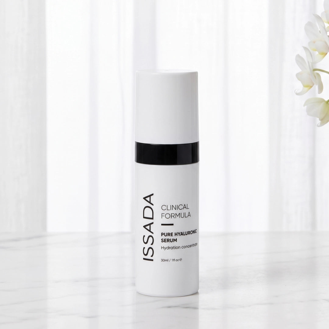 ISSADA Pure Hyaluronic Serum bottle displayed on a white marble surface with soft curtains and orchid blooms.