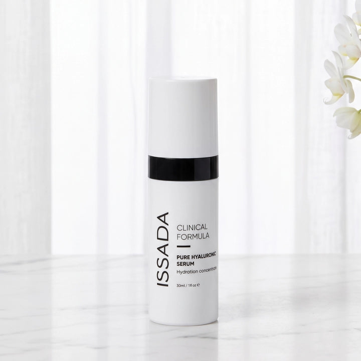ISSADA Pure Hyaluronic Serum bottle displayed on a white marble surface with soft curtains and orchid blooms.