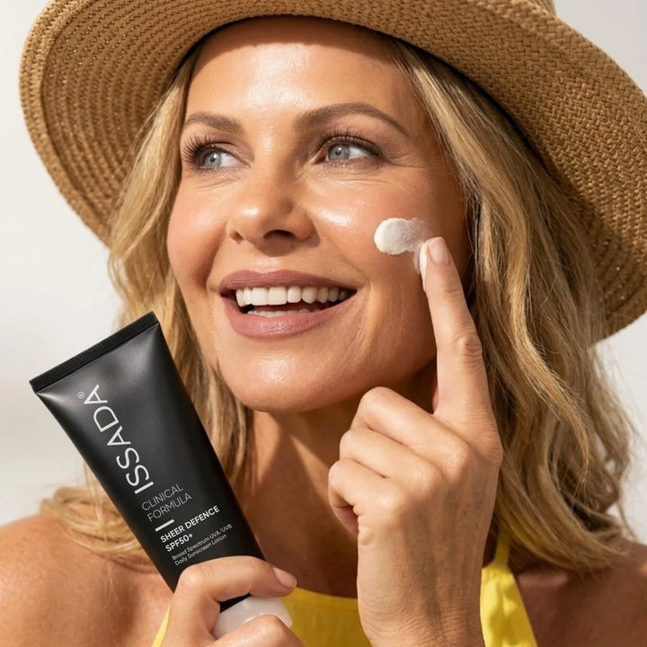 Smiling woman in a sun hat applying ISSADA Sheer Defence SPF50+ sunscreen to her cheek while holding the tube.