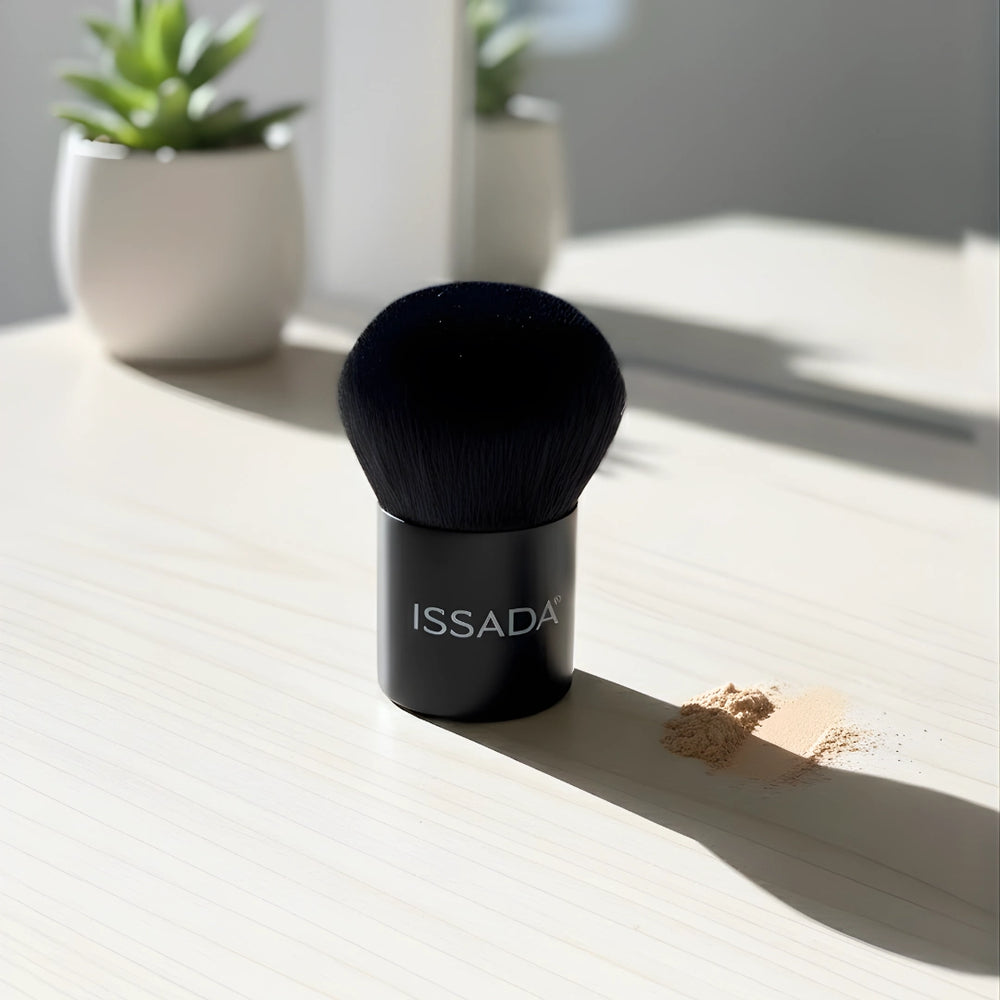 ISSADA small vegan kabuki makeup brush with dense black bristles on a sunlit vanity next to loose powder