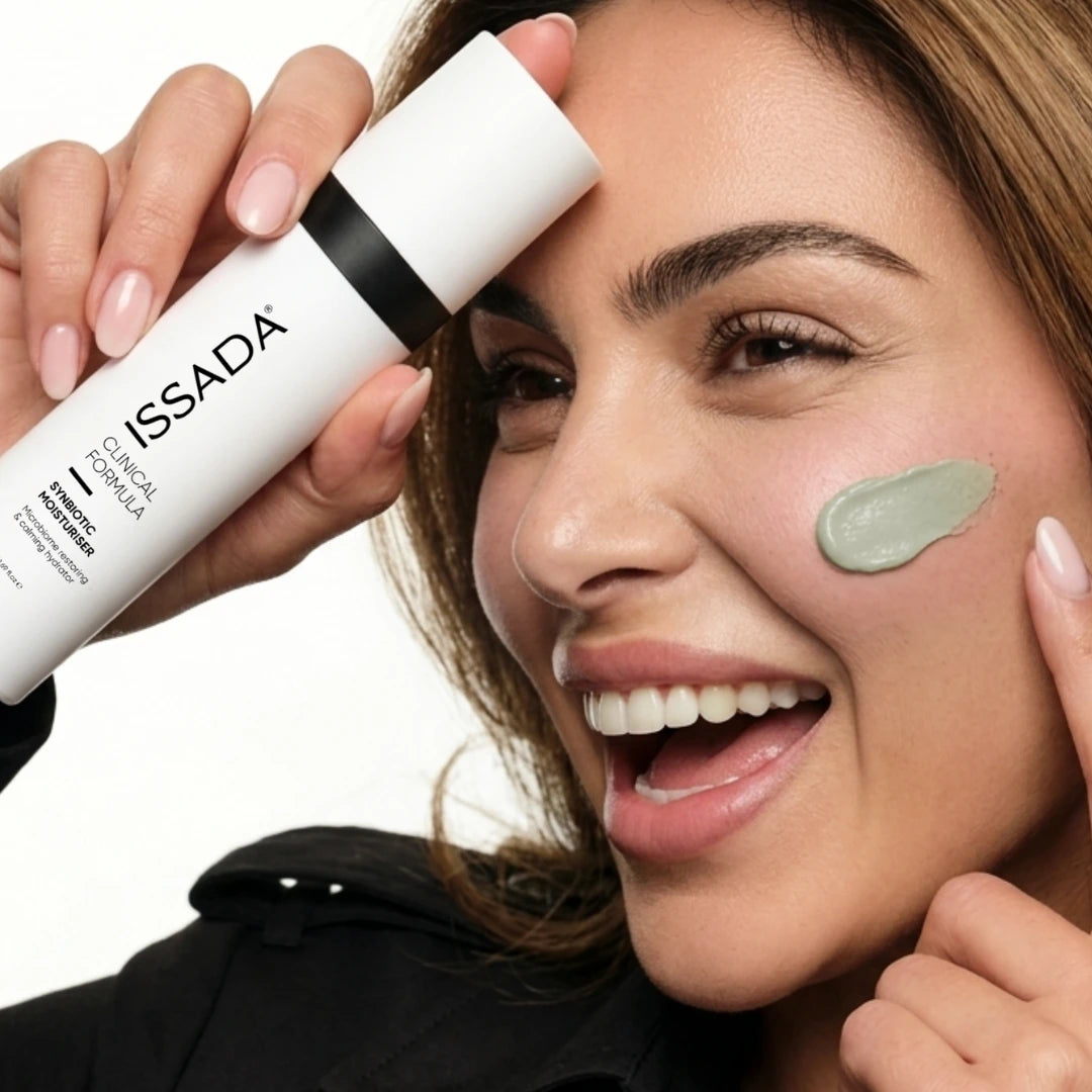 Smiling woman holding ISSADA Synbiotic Moisturiser with a green cream swatch on her cheek against a white background.
